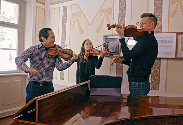 3 people playing violins around a piano
